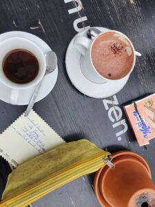 Auf einem schwarzen Tisch stehen eine heiße Schokolade und ein Kaffee. Danben liegt eine offene Packung Mannerschnitten, und die Postkarte mit dem Hinweistext. Der Adressbereich der Postkarte wird durch ein gelbes Federpennal verdeckt.