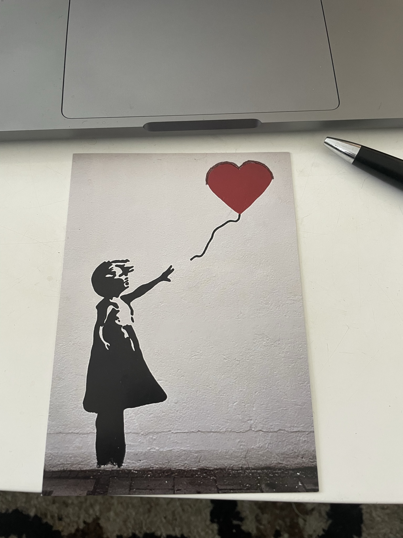 Eine Postkarte mit dem Banksy-Graffiti eines kleinen Kindes, das einen roten herzförmigen Luftballon fliegen lässt. Die Rundungen des Luftballons wurden mit schwarzem Kugelschreiber nachgezogen.