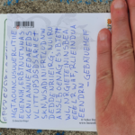 Foto einer Postkarte, beschriftet mit Buchstabensalat. An einem Strichcode wurde eine Ziffer "1" rot eingekreist. Der Adressbereich wird von einer Hand verdeckt.